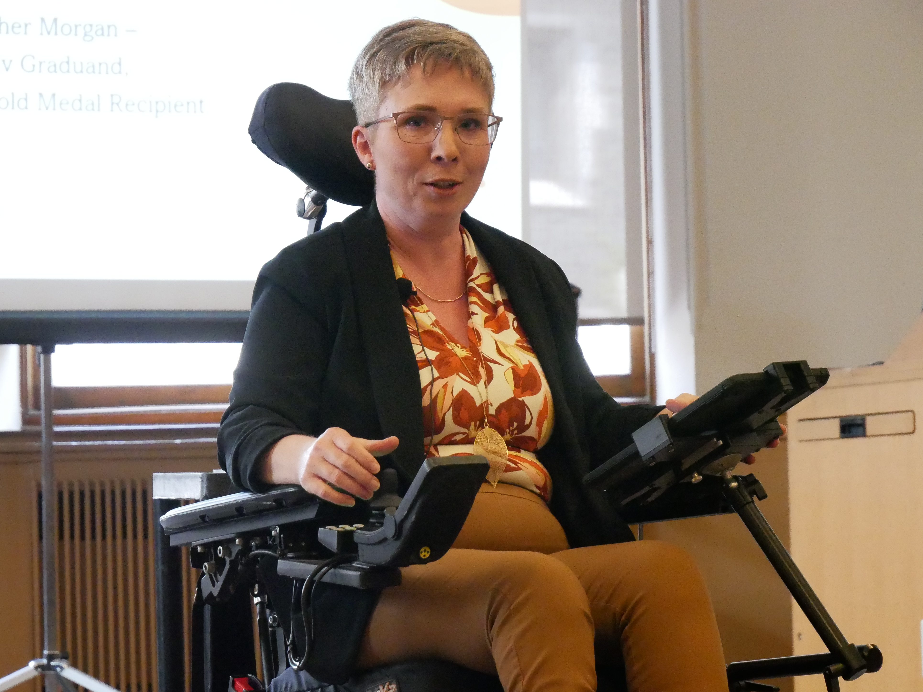 Heather sits in her power wheelchair wearing mustard yellow trousers, a multicolour top and a black suit jacket. A powerpoint slide is visible behind her wheelchair. She uses an ipad attached to her chair for accessing her notes, and she faces toward the camera with a look of gentle engagement.