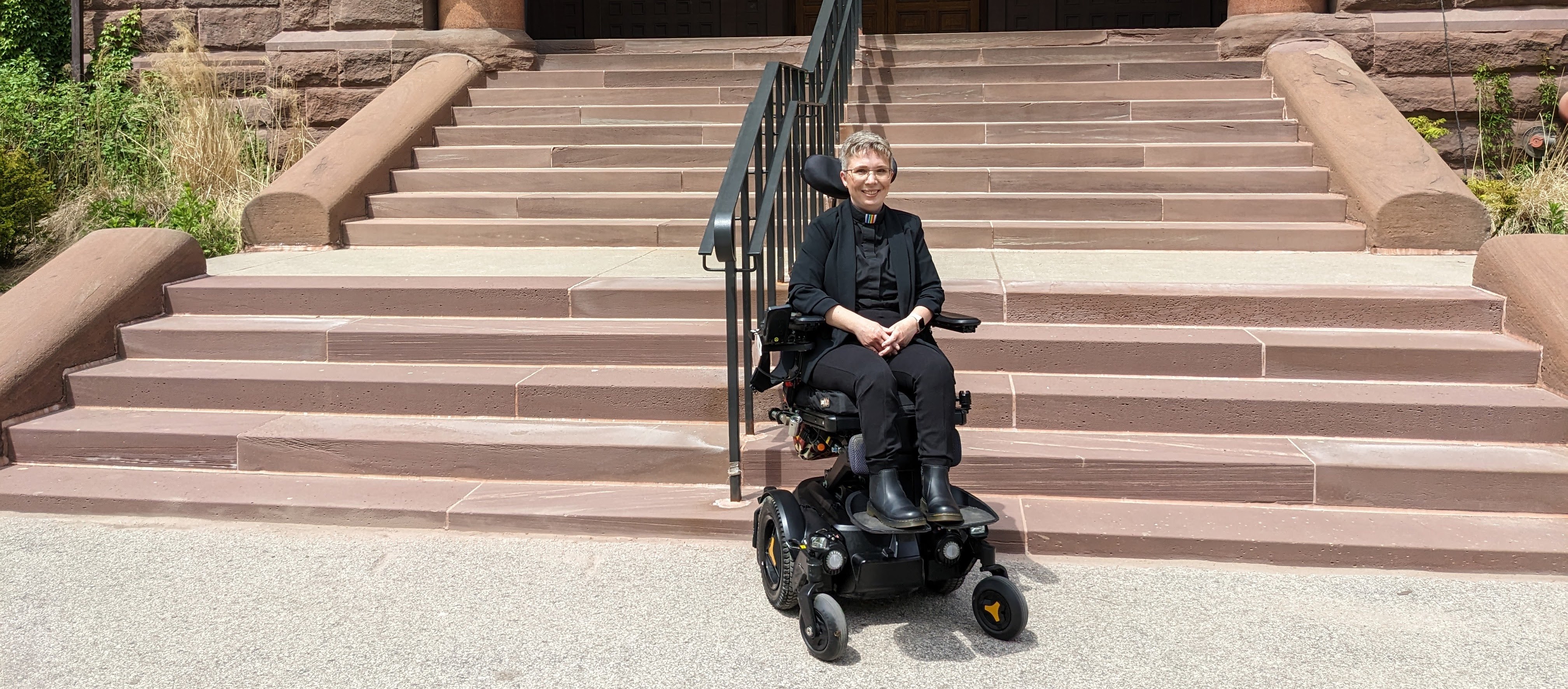 Heather sits in her elevated power wheelchair in front of a flight of steps in a red stone. She is dressed all in black apart from her rainbow clergy collar.